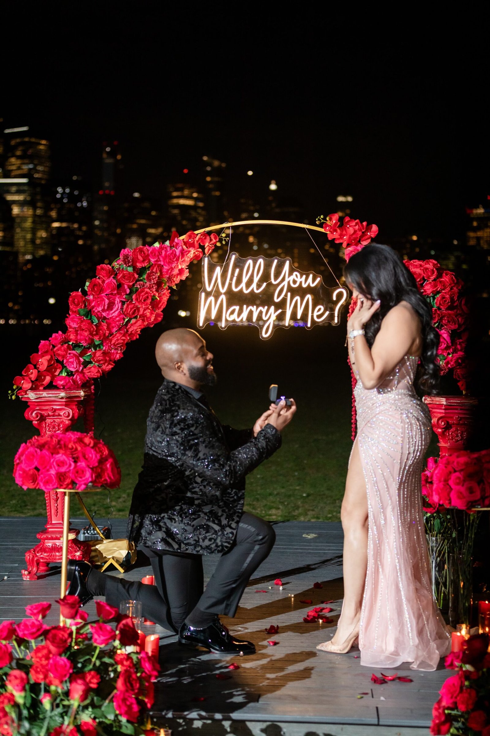 Partner on one knee beneath a red rose arch lit by a Will You Marry Me sign